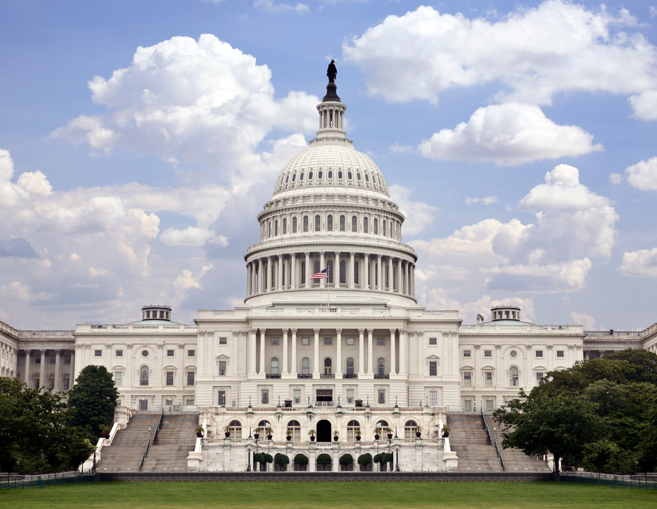 The United States Capitol building in Washington, D.C., with its iconic white dome topped by a statue, flanked by long wings, and fronted by steps and green lawn under a partly cloudy sky.