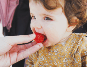 A baby takes a bite of a strawberry held by an adult, showing early exposure to fruits as solid foods.