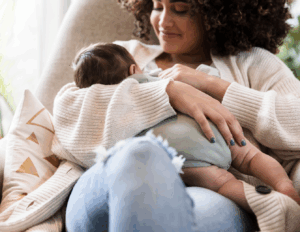 A caregiver sits in a chair holding and feeding an infant, emphasizing closeness and early nourishment during infancy.