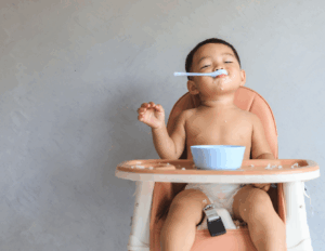 A toddler sits in a high chair feeding themself from a bowl, with food on their face and hands.
