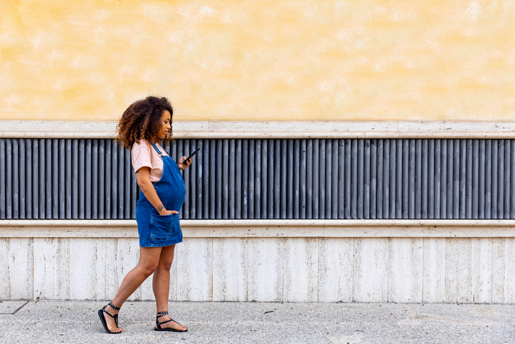 A pregnant person walks along a sidewalk while holding a phone, wearing a casual outfit in an urban setting.