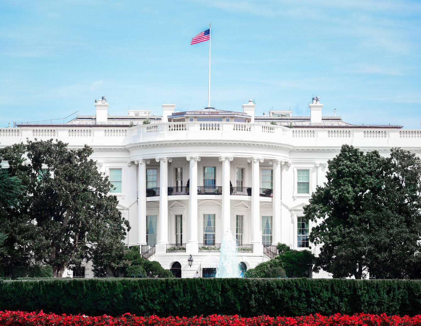 The White House in Washington, D.C., with an American flag flying above it and a fountain and trees in the foreground, representing the federal administration behind proposed budget decisions impacting programs for young children and families.