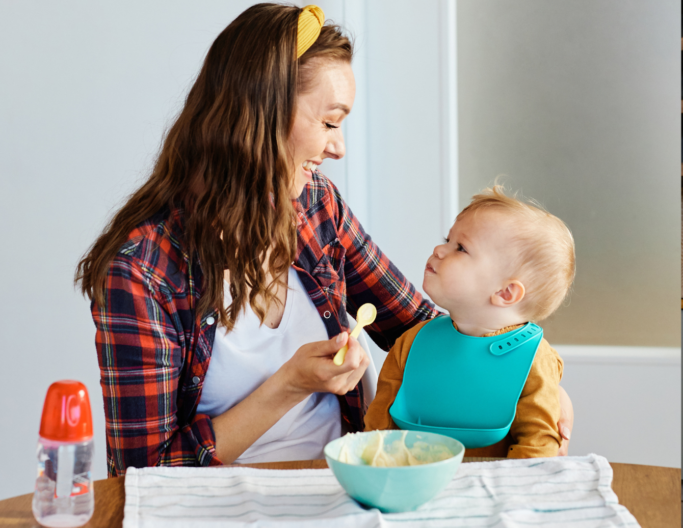 A smiling caregiver sits at a table feeding a toddler wearing a blue bib. A bowl of food and baby bottle are on the table during mealtime at home.