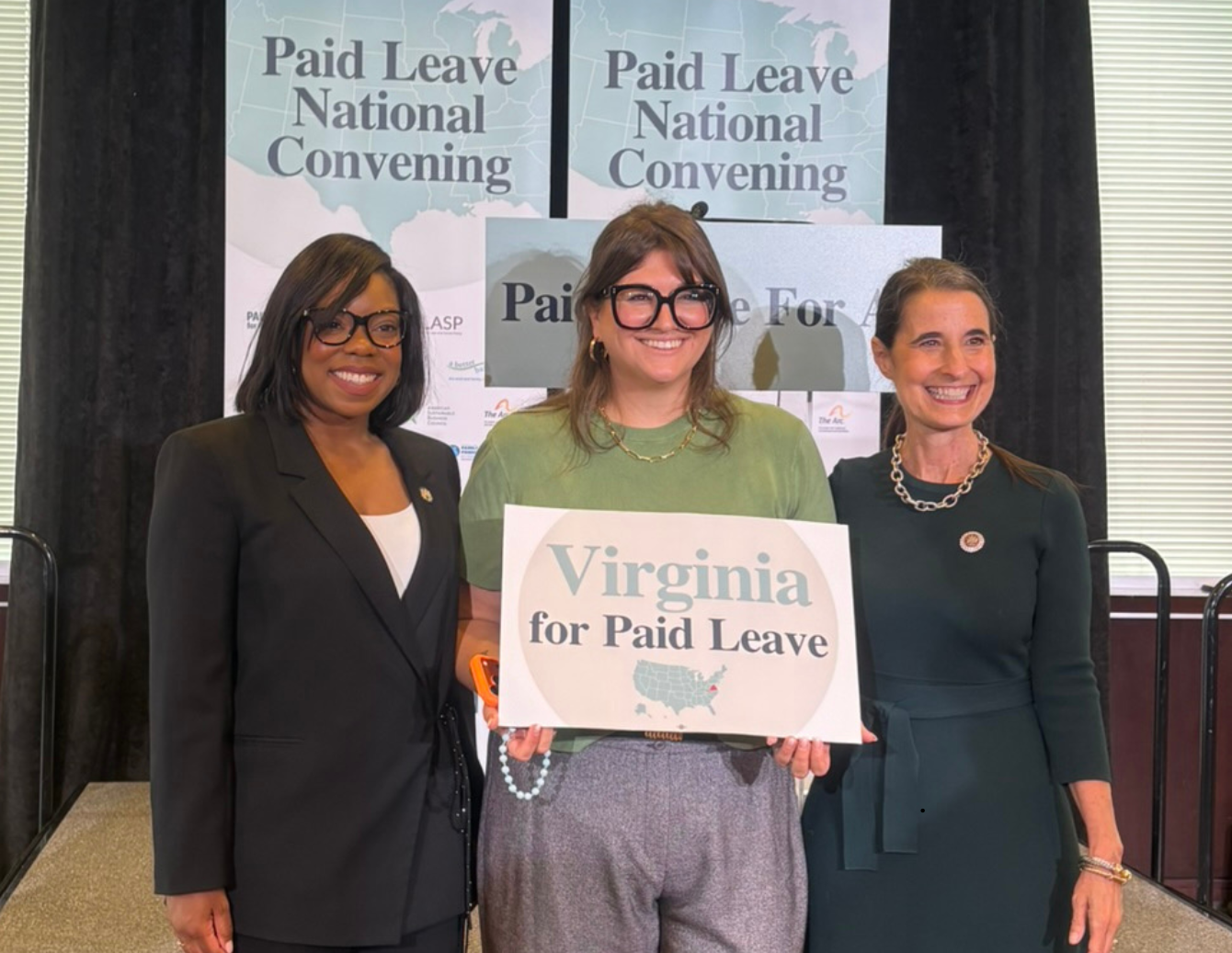 Three event participants stand in front of “Paid Leave National Convening” signage, holding a sign that reads “Virginia for Paid Leave.”