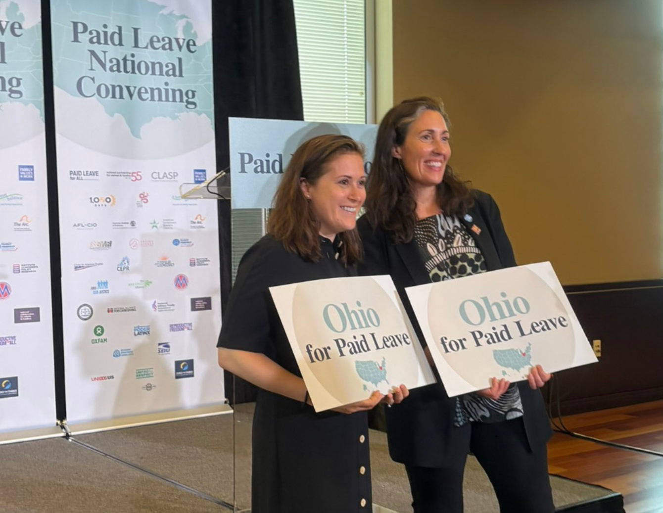Two event participants stand on a stage at the Paid Leave National Convening holding “Ohio for Paid Leave” signs in front of branded banners in Washington, D.C.