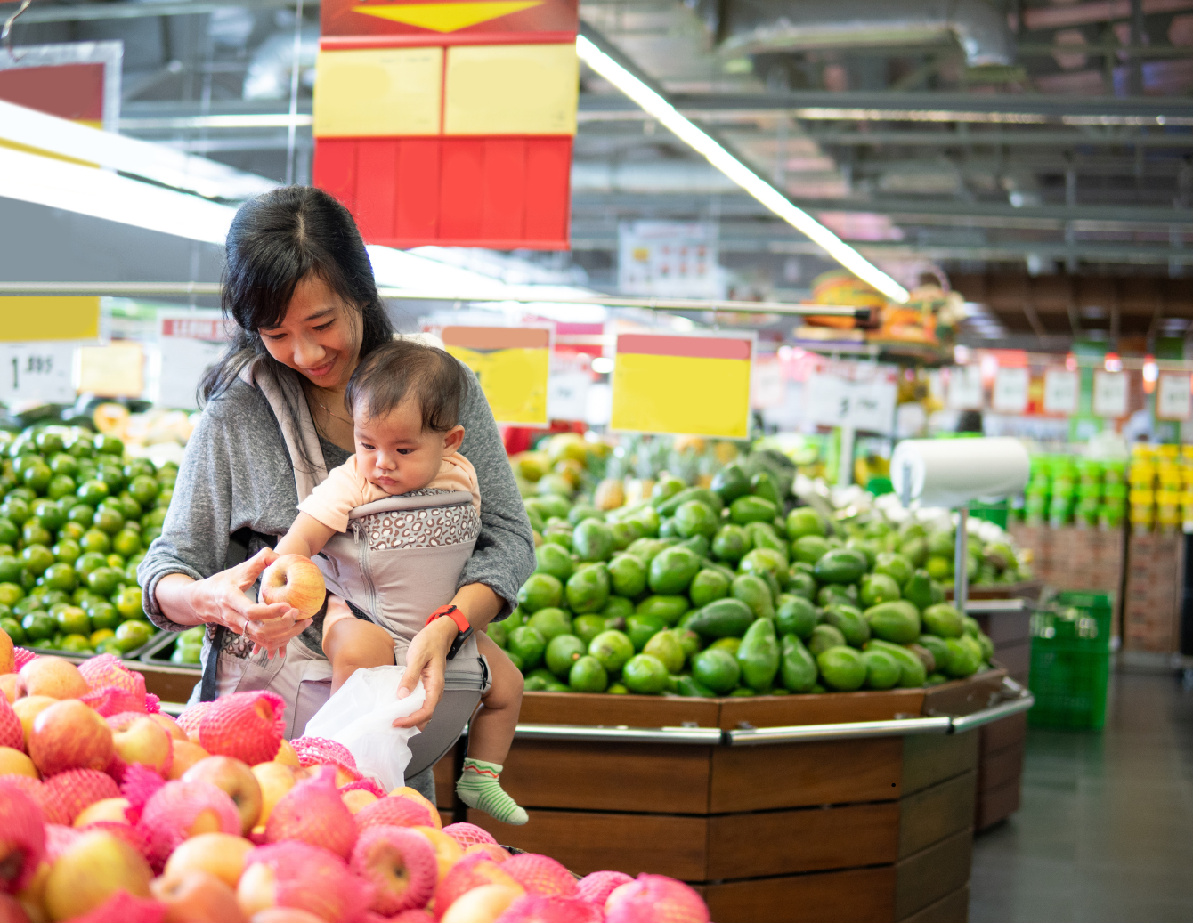 A woman wearing a baby carrier shops in the produce section of a grocery store while holding an apple. Fresh fruits and green vegetables are displayed around her as she shops with her young child.