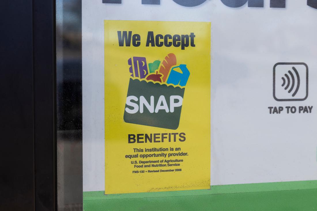 Close-up of a yellow “We Accept SNAP Benefits” sign with grocery icons on a storefront window, next to a tap-to-pay symbol, indicating the location accepts food assistance benefits.