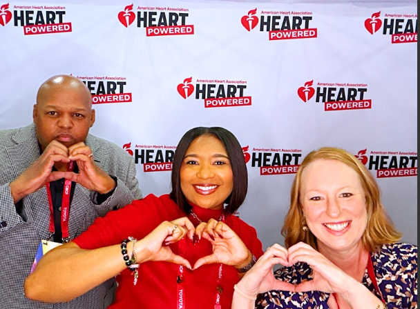 Three people pose in front of an American Heart Association Heart Powered backdrop, making heart shapes with their hands at a CBC conference booth.